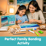 A woman and a young girl are sitting together at a wooden table, working on a sand and shell studio craft kit that forms a blue ocean wave design. The kit includes various shells, sand, paint tubes, and a box labeled 'Sand and Shell Studio'.